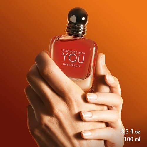 Hands holding a bottle of Stronger With You Intensely perfume against an orange background.